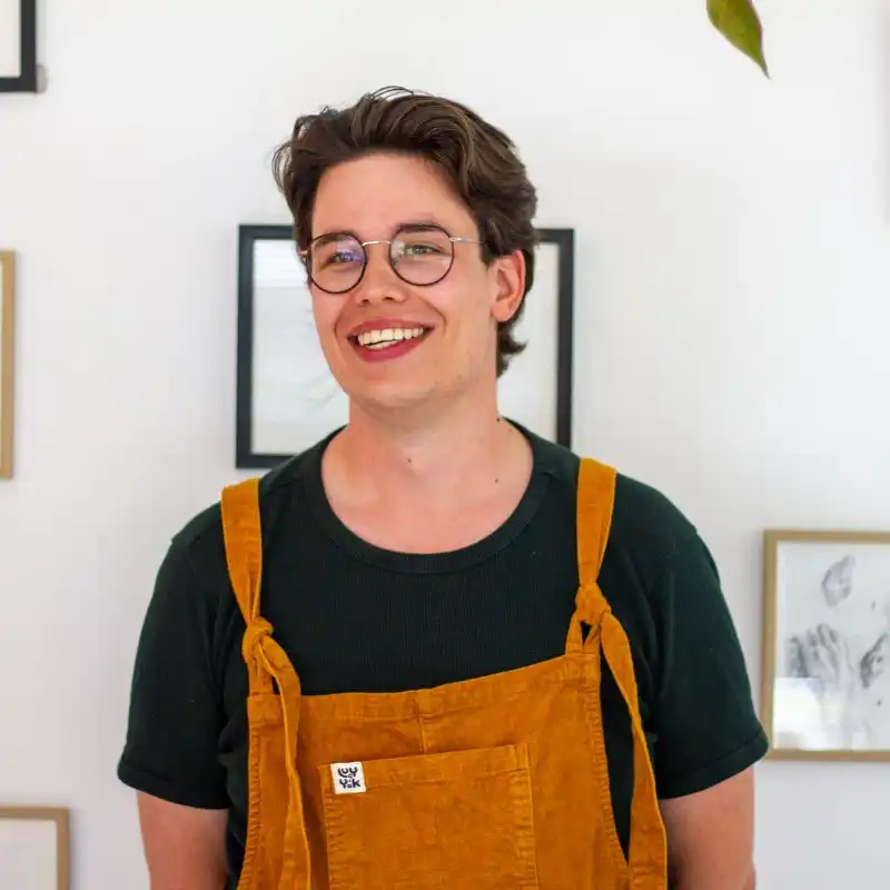 Tijn is een man met middenlang bruin haar. Hij draagt een bril, een groen shirt met daarover een gele tuinbroek. Hij lacht naar de camera.